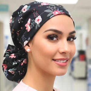Puede incluir: Un gorro quirúrgico negro con un estampado floral de flores rosas y blancas y hojas verdes. El gorro está atado en un moño en la parte posterior. La persona lleva una camiseta quirúrgica rosa y un pendiente de perla.