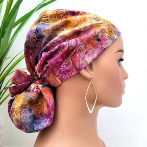 Peut inclure: Un couvre-chef coloré avec des motifs roses, oranges et bleus. Le tissu est noué à l'arrière, formant une sorte de chignon. Le couvre-chef est porté avec des boucles d'oreilles dorées en forme de losange.