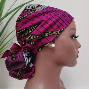 May include: A magenta head wrap with a geometric pattern of black, yellow, and white lines. The head wrap is tied in a bow at the back. The head wrap is worn on a mannequin head.