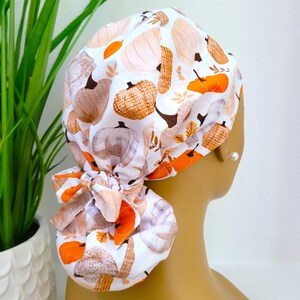 Pode incluir: Uma touca cirúrgica branca com um padrão repetido de abóboras em tons de laranja, castanho e bege. A touca tem as costas franzidas e um laço a condizer. O design evoca o outono e os temas da colheita.