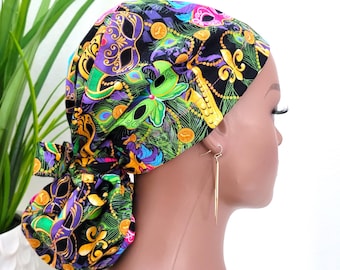 May include: A black scrub hat with a colorful Mardi Gras print featuring masks, beads, and a fleur-de-lis. The hat is tied at the back with a bow.
