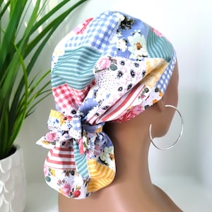 May include: A colorful patchwork scrub hat with a bow. The hat features various patterns, including gingham, stripes, and floral prints in shades of blue, red, yellow, and white. The bow is tied at the back. A silver hoop earring is visible.