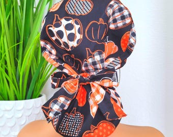 Pode incluir: Uma touca cirúrgica preta com um padrão de abóbora em laranja, branco e preto. As abóboras têm vários desenhos, incluindo listras, bolinhas e xadrez. A touca tem um laço a condizer na parte de trás.