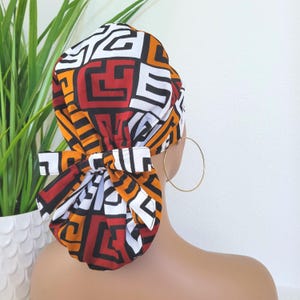 May include: A colorful head wrap featuring a geometric pattern in red, black, white, and orange. The head wrap is tied in a bow at the back. A gold hoop earring is visible. The background includes a white wall and a green plant.