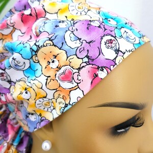 May include: A scrub cap featuring a colorful, repeating pattern of cartoon bears in shades of pink, purple, yellow, and blue. The bears are depicted with various symbols on their chests, including hearts, moons, and suns, set against a white background.