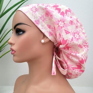 May include: A pink and white patterned surgical scrub cap with a geometric design. The cap is tied at the back with matching fabric ties. The cap is on a mannequin head.