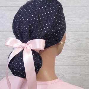 May include: A navy blue scrub cap featuring a white polka dot pattern, designed with a gathered back to accommodate hair. The cap is accented with a light pink satin ribbon tied into a bow at the base, providing a secure and adjustable fit.