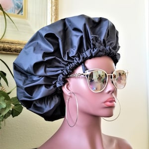 Peut inclure: Un bonnet de nuit en satin noir avec un sommet plissé. Le bonnet est porté par un mannequin avec de grandes boucles d'oreilles créoles et des lunettes de soleil à monture dorée.