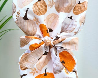 Pode incluir: Uma touca cirúrgica branca com um padrão repetido de abóboras em tons de laranja, castanho e bege. A touca é amarrada com um laço a condizer. O design inclui folhas de outono e um padrão xadrez.