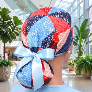 Può includere: Una cuffia chirurgica con un motivo patchwork di quadrati di tessuto rosso, blu scuro e azzurro con piccole stampe floreali bianche. La cuffia è fissata sul retro con un ampio nastro di raso azzurro annodato a fiocco decorativo.