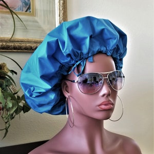 May include: A bright blue satin shower cap with a gathered top and a tie closure. The cap is worn on a mannequin with large hoop earrings and aviator sunglasses.