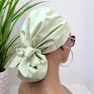 May include: A light green surgical scrub hat with a bow tied at the back. The hat is made of a soft, breathable fabric and is designed to be comfortable to wear for long periods of time.