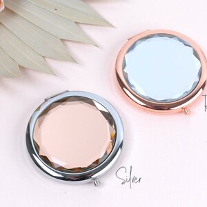 May include: Two compact mirrors with a faceted design. One mirror has a silver frame and a pink glass, the other has a rose gold frame and a clear glass. The text "Rose Gold" is written below the rose gold mirror and "Silver" is written below the silver mirror.