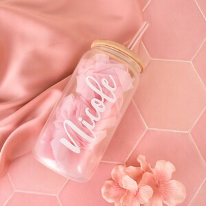 Puede incluir: Un tarro de vidrio transparente con tapa de bamb&uacute; y una pajita transparente. El tarro est&aacute; lleno de papel de seda rosa y tiene el nombre "Nicole" escrito en letras blancas. El tarro est&aacute; sobre una superficie de azulejos rosa.