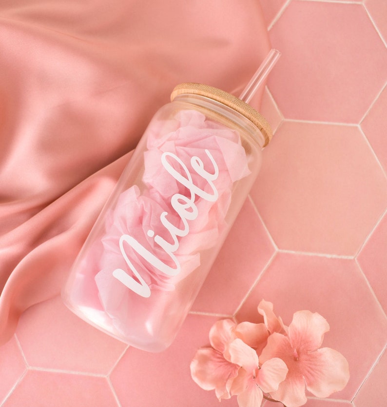 Puede incluir: Un tarro de vidrio transparente con tapa de bamb&uacute; y una pajita transparente. El tarro est&aacute; lleno de papel de seda rosa y tiene el nombre "Nicole" escrito en letras blancas. El tarro est&aacute; sobre una superficie de azulejos rosa.