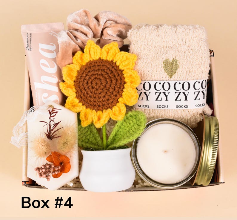 May include: A gift box filled with self-care items. Includes a crochet sunflower, a candle in a jar, a tube of shea lotion, a pair of socks, a scrunchie, and a decorative item with dried flowers. The box is labeled "Box #4".