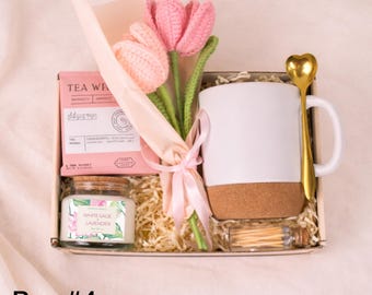 May include: A gift box with a white mug, a gold heart-shaped spoon, a bouquet of pink crochet tulips, a candle, tea, and matches. The candle is labeled "White Sage Lavender". The box is filled with shredded paper.