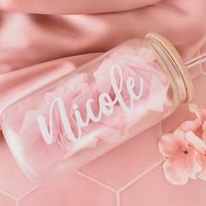 Puede incluir: Una botella de agua de vidrio transparente con tapa de bamb&uacute; y una pajita. La botella tiene el nombre "Nicole" escrito en ella con letras blancas. La botella est&aacute; sobre una superficie de azulejos rosa con flores rosas.