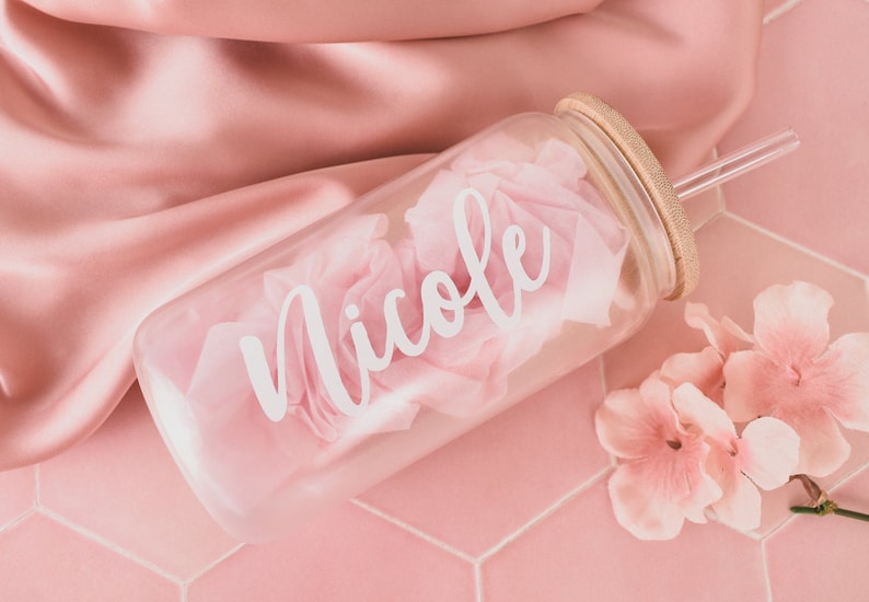 Puede incluir: Una botella de agua de vidrio transparente con tapa de bamb&uacute; y una pajita. La botella tiene el nombre "Nicole" escrito en ella con letras blancas. La botella est&aacute; sobre una superficie de azulejos rosa con flores rosas.