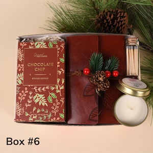 May include: A gift box containing a leather-bound journal, a box of chocolate chip cookies, a small candle, and a vial of matches. The journal is decorated with a pinecone and berries. The box is labeled "Box #6".