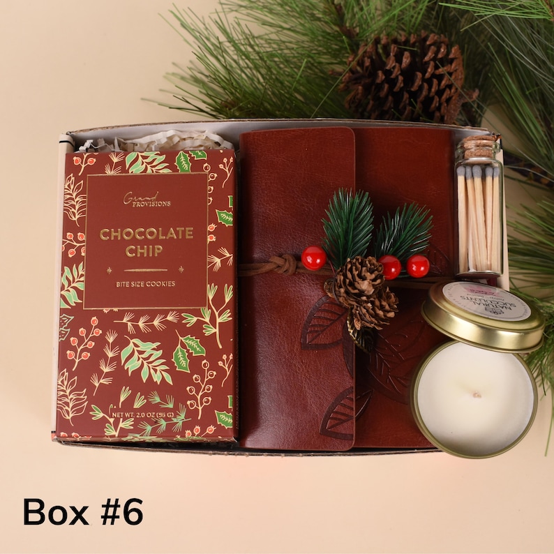 May include: A gift box containing a leather-bound journal, a box of chocolate chip cookies, a small candle, and a vial of matches. The journal is decorated with a pinecone and berries. The box is labeled "Box #6".