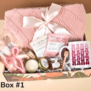 Puede incluir: Una caja de regalo rosa y blanca llena de varios art&iacute;culos, incluyendo una manta rosa, una taza con el texto "MAMA" repetido en un degradado de rosa, un tarro de miel, una vela y un paquete de t&eacute;. La caja est&aacute; decorada con un patr&oacute;n de hojas tropicales.