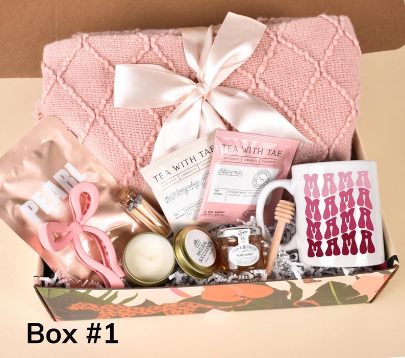 Puede incluir: Una caja de regalo rosa y blanca llena de varios art&iacute;culos, incluyendo una manta rosa, una taza con el texto "MAMA" repetido en un degradado de rosa, un tarro de miel, una vela y un paquete de t&eacute;. La caja est&aacute; decorada con un patr&oacute;n de hojas tropicales.