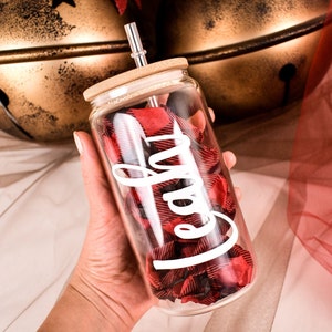 Puede incluir: Un vaso de vidrio transparente con tapa de bamb&uacute; y pajita plateada. El vaso est&aacute; lleno de tela a cuadros roja y negra y la palabra "Cafe" en escritura blanca. El vaso se sostiene en una mano.