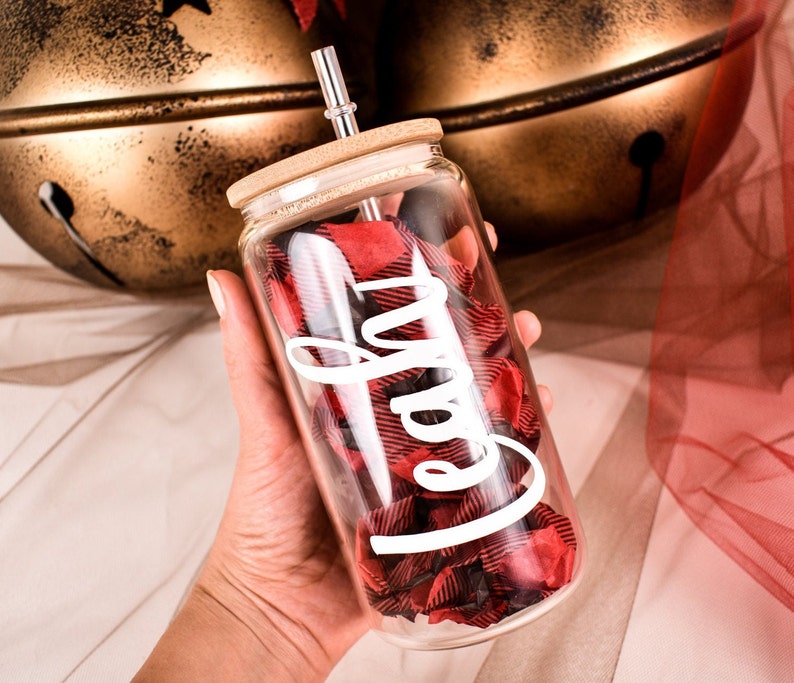 Puede incluir: Un vaso de vidrio transparente con tapa de bamb&uacute; y pajita plateada. El vaso est&aacute; lleno de tela a cuadros roja y negra y la palabra "Cafe" en escritura blanca. El vaso se sostiene en una mano.