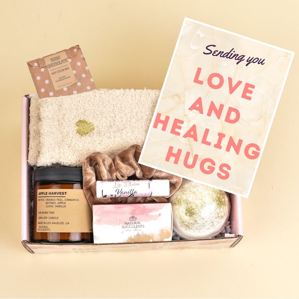 May include: A pink gift box filled with self-care items, including a candle, bath bomb, lip balm, and a cozy white blanket. The box also includes a card that reads "Sending you LOVE AND HEALING HUGS".