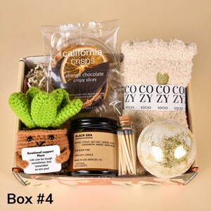 May include: A gift box containing an assortment of items. Includes orange chocolate crispy slices, a crochet succulent, a black sea scented candle, a bottle of matches, a bath bomb, and cozy socks. The box is labeled "Box #4".