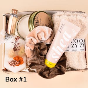May include: A gift box labeled "Box #1" filled with self-care items. Includes a candle, matches, a tube of shea butter lotion, fuzzy socks, and velvet scrunchies in shades of beige and brown. The box is on a light beige background.