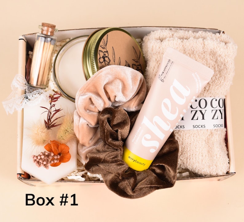 May include: A gift box labeled "Box #1" filled with self-care items. Includes a candle, matches, a tube of shea butter lotion, fuzzy socks, and velvet scrunchies in shades of beige and brown. The box is on a light beige background.