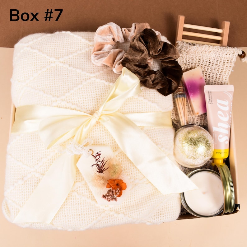 May include: A gift box labeled "Box #7" filled with self-care items. It includes a cream-colored knitted blanket tied with a yellow ribbon, two velvet scrunchies, a candle, bath bomb, soap, and lotion. A tag with dried flowers is attached.