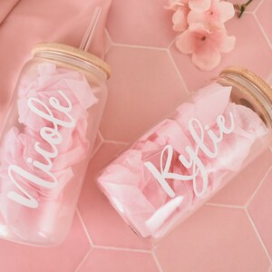 Puede incluir: Dos vasos de vidrio transparentes con tapas de bamb&uacute; y pajitas transparentes. Los vasos est&aacute;n llenos de papel de seda rosa y tienen los nombres "Nicole" y "Kylie" escritos en blanco.