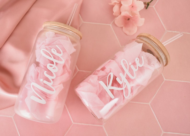 Puede incluir: Dos vasos de vidrio transparentes con tapas de bamb&uacute; y pajitas transparentes. Los vasos est&aacute;n llenos de papel de seda rosa y tienen los nombres "Nicole" y "Kylie" escritos en blanco.