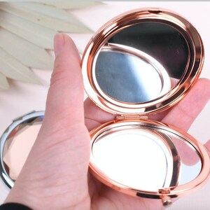 May include: A rose gold compact mirror with a hinged lid. The mirror is open and the reflection of the hand holding the mirror is visible.