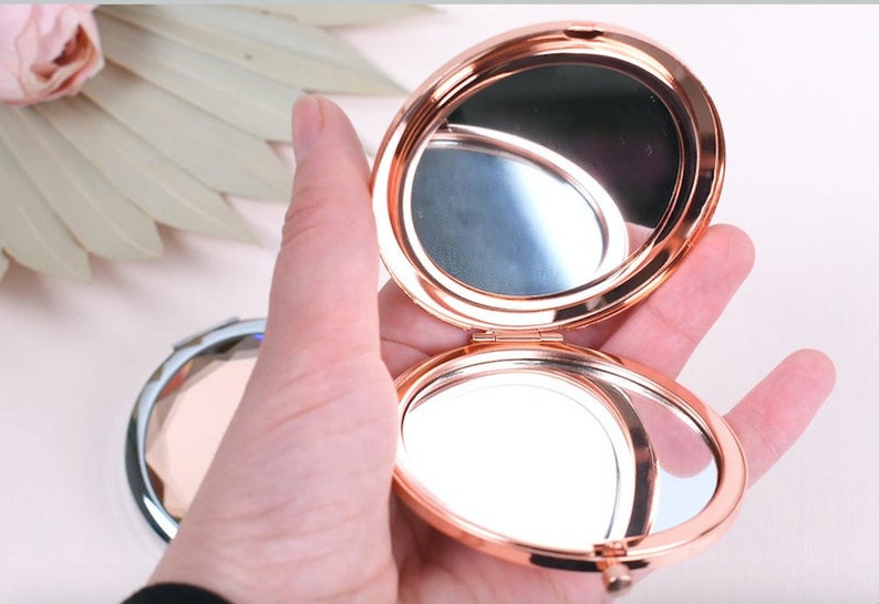 May include: A rose gold compact mirror with a hinged lid. The mirror is open and the reflection of the hand holding the mirror is visible.