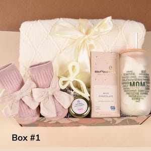 May include: A gift box containing baby items and treats. Includes pink baby booties with bows, a cream-colored blanket, a chocolate bar, a candle, and a jar of succulents. The box is labeled "Box #1".