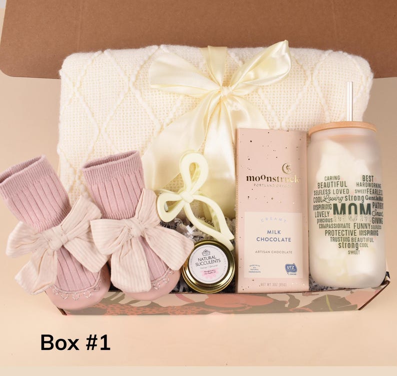May include: A gift box containing baby items and treats. Includes pink baby booties with bows, a cream-colored blanket, a chocolate bar, a candle, and a jar of succulents. The box is labeled "Box #1".