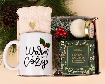 May include: A gift box with a white mug that says "Warm and Cozy" with holly accents, a pair of cream-colored socks, a candle, and a box of Sea Salt Caramel chocolate cookies. The box is labeled "Box #6".