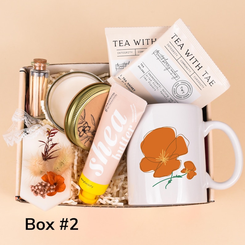 May include: A gift box filled with self-care items. It includes a white mug with orange floral design, tea sachets, a shea butter tube, a candle, matches, and a decorative sachet. The box is labeled "Box #2".