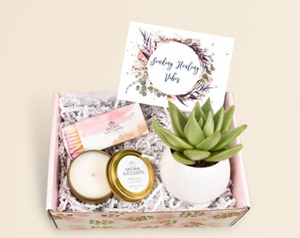 May include: A gift box filled with a small succulent plant in a white pot, a candle in a gold tin, a box of matches, and a card that says "Sending Healing Vibes".