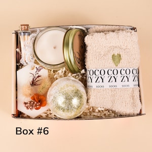 May include: A gift box containing a candle in a glass jar, a bath bomb, a pair of socks, a decorative tag, and a small bottle of matches. The socks are beige with a green heart. The box is labeled "Box #6".