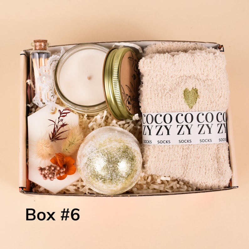 May include: A gift box containing a candle in a glass jar, a bath bomb, a pair of socks, a decorative tag, and a small bottle of matches. The socks are beige with a green heart. The box is labeled "Box #6".