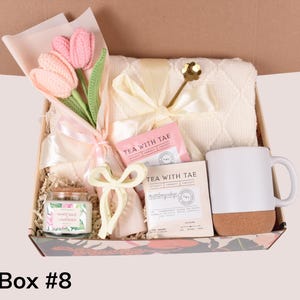 May include: A gift box filled with a bouquet of pink crochet tulips, a white mug with a cork base, a white blanket, tea sachets, a candle, and a decorative bow. The box is decorated with a floral pattern.