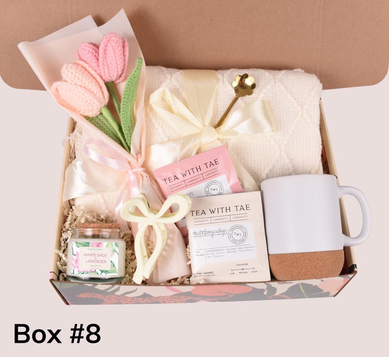 May include: A gift box filled with a bouquet of pink crochet tulips, a white mug with a cork base, a white blanket, tea sachets, a candle, and a decorative bow. The box is decorated with a floral pattern.