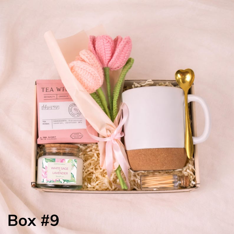 May include: A gift box with a white mug, gold heart-shaped spoon, pink knitted tulips, tea, a candle, and matches. The candle is labeled "White Sage Lavender". The box is filled with shredded paper.