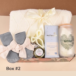 May include: A gift box containing a cream-colored baby blanket, gray baby socks with pink bows, a jar of dark chocolate almond, a glass tumbler with the word "MOM" and other words, and a jar of natural succulents. The box is labeled "Box #2".