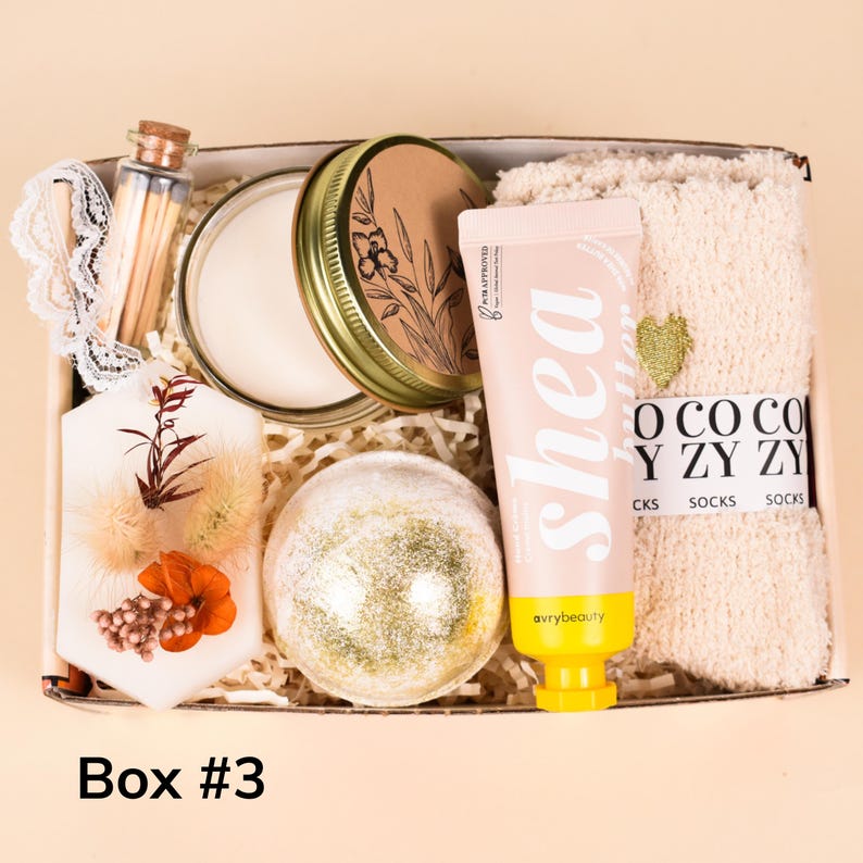 May include: A gift box filled with self-care items. Includes a candle, bath bomb, shea lotion, cozy socks, matches, and a decorative lace piece. The box is labeled "Box #3".